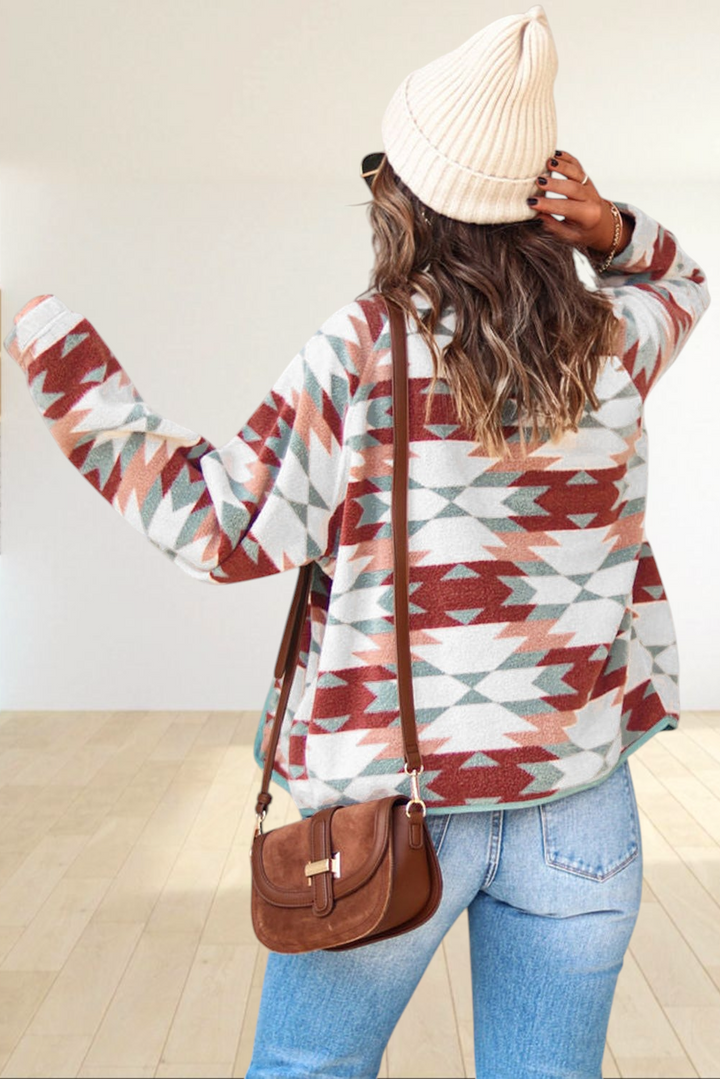 Baya Wear geometric Aztec fleece jacket in red, cream, and blue worn with jeans, a beanie, and a brown crossbody bag.