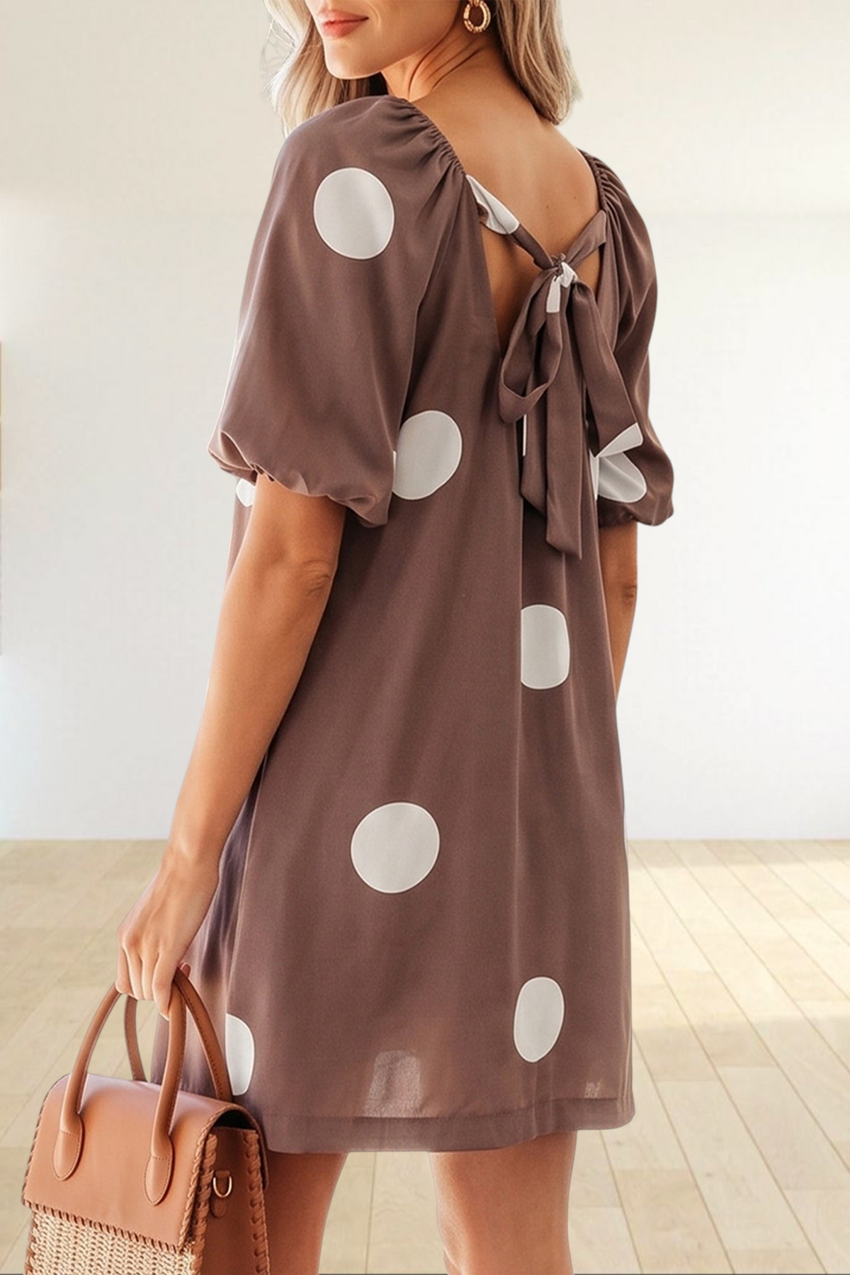 Baya Wear brown mini dress with white polka dots, puff sleeves, and a knot-back detail worn by a woman by a pool.