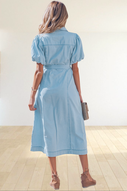 Baya Wear light blue denim midi dress with puff sleeves and tie-waist belt, worn by a woman on a sunny sidewalk.
