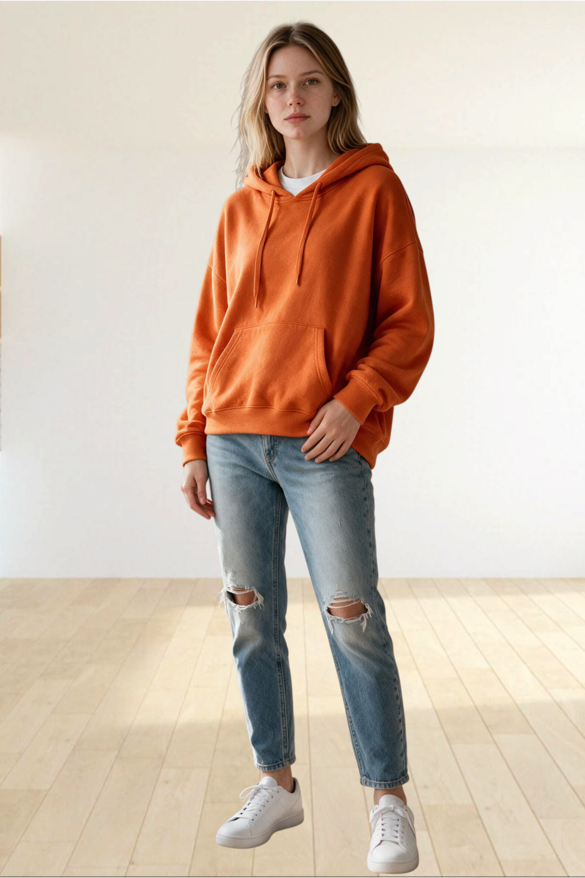 A woman wearing a Baya Wear oversized orange vintage washed pullover hoodie with jeans in a bright minimalist room.